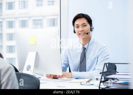 Sorridente bell'uomo asiatico che indossa cuffie con microfono che funzionano durante la chiamata centrale come telemarketer Foto Stock
