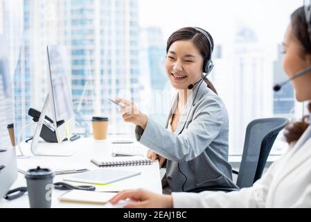 Sorridente bella donna asiatica telemarketing agente di servizio di cliente che lavora dentro ufficio call center con un collega Foto Stock