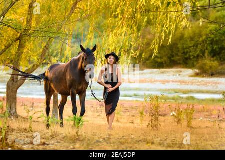 Una ragazza in un bel vestito nero e un nero hat cammina con un cavallo attraverso il campo Foto Stock