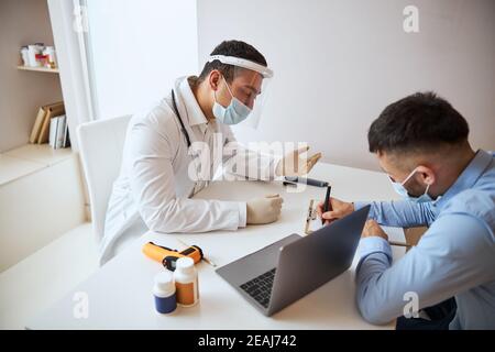 Terapista professionista che parla con l'uomo in camicia blu mentre scrive qualcosa vicino al medico Foto Stock