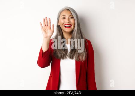 Amichevole anziana signora asiatica in blazer elegante che dice hi, muovendo la mano e salutarvi con sorriso felice, in piedi su sfondo bianco Foto Stock