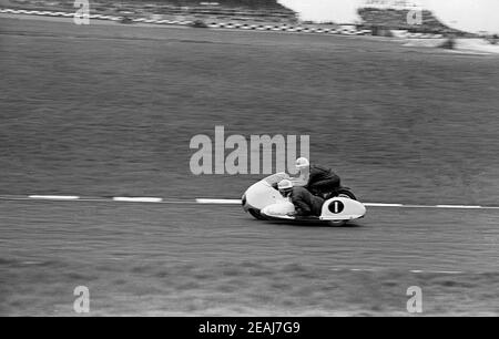 Anni '60, storico, motorsport, corse sidecar, un motociclista e un passeggero sidecar sul loro veicolo - numerato uno - sulla pista di gara, Inghilterra, Regno Unito. Il sidecar racing è uno sport che utilizza una moto a 3 ruote su misura ed è l'unica forma di motorsport in cui sia il passeggero che il conducente guidano il veicolo. Il veicolo viaggia ad alta velocità e lo sport è emozionante da guardare. Sia il pilota che il passeggero indossano i tradizionali abiti in pelle nera dell'epoca. Foto Stock