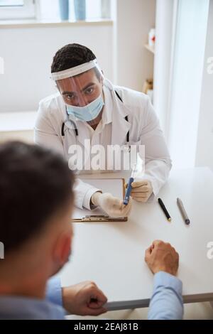 Medico sicuro di sé in uniforme bianca e maschera protettiva seduta alla scrivania con l'attrezzo speciale in mano Foto Stock