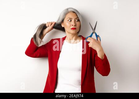 Donna asiatica indecisa che tiene forbici e che guarda dubbioso, pensando di tagliare i capelli, cambiando il taglio dei capelli, in piedi su sfondo bianco Foto Stock