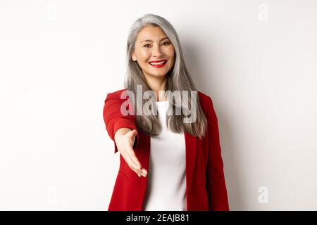 Donna d'affari asiatica professionista con i capelli grigi, salutando, allunga la mano per la stretta di mano e sorridendo, in piedi su sfondo bianco Foto Stock