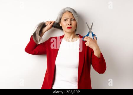 Donna asiatica indecisa che tiene forbici e che guarda dubbioso, pensando di tagliare i capelli, cambiando il taglio dei capelli, in piedi su sfondo bianco Foto Stock