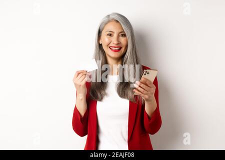 Donna asiatica di mezza età che usa carta di credito e smartphone di plastica per fare acquisti online, in piedi su sfondo bianco Foto Stock