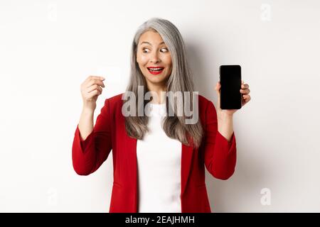 Allegra donna asiatica matura che mostra schermo vuoto smartphone e carta di credito, concetto di e-commerce Foto Stock