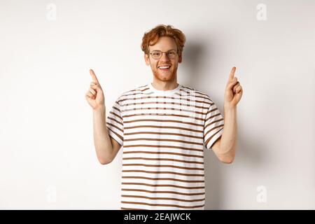 Divertente giovane ragazzo rosso in occhiali nerdy puntando le dita lateralmente, mostrando due offerte promo, in piedi felice su sfondo bianco Foto Stock