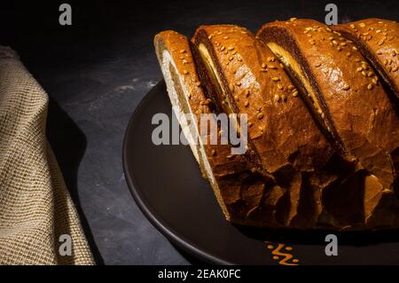 Insolito pane fresco a due colori in un piatto di ceramica e un tovagliolo intrecciato su uno sfondo nero, foto in un tasto basso in luce dura, primo piano Foto Stock