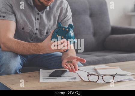 L'uomo seduto ad un tavolo pieno di fatture non pagate scuote fuori l'ultimo centesimo dalla banca piggy. Spendi i tuoi ultimi risparmi. Disoccupazione, povertà, concetto fallimentare. Foto Stock