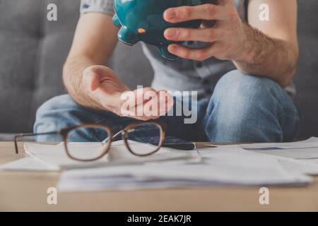 L'uomo seduto ad un tavolo pieno di fatture non pagate scuote fuori l'ultimo centesimo dalla banca piggy. Spendi i tuoi ultimi risparmi. Disoccupazione, povertà, concetto fallimentare. Foto Stock