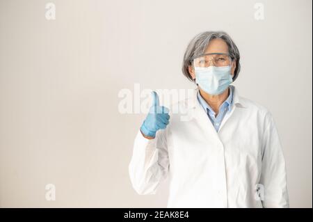 Senior femmina medico in accappatoio medico, occhiali di protezione, maschera facciale, e guanti in lattice, in posizione isolata su sfondo grigio, mostrando pollice-up, completamente Foto Stock