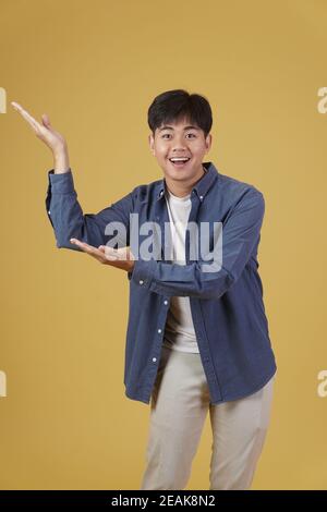 ritratto di felice bel giovane asiatico vestito casualmente presentando con gesto palmo aperto al copyspace. sfondo giallo studio Foto Stock