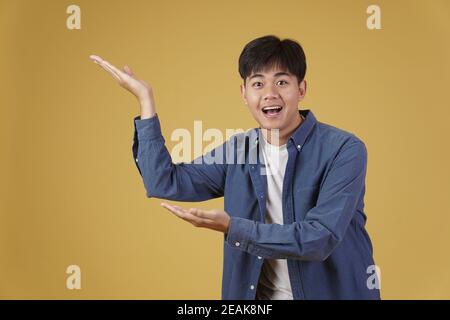 ritratto di felice bel giovane asiatico vestito casualmente presentando con gesto palmo aperto al copyspace. sfondo giallo studio Foto Stock