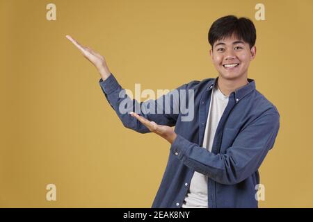 ritratto di felice bel giovane asiatico vestito casualmente presentando con gesto palmo aperto al copyspace. sfondo giallo studio Foto Stock