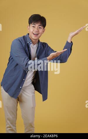 ritratto di felice bel giovane asiatico vestito casualmente presentando con gesto palmo aperto al copyspace. sfondo giallo studio Foto Stock