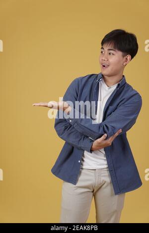 ritratto di felice bel giovane asiatico vestito casualmente presentante con gesto aperto della mano palma al copyspace isolato su giallo sfondo studio Foto Stock