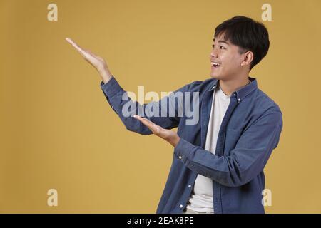 ritratto di felice bel giovane asiatico vestito casualmente presentando con gesto palmo aperto al copyspace. sfondo giallo studio Foto Stock