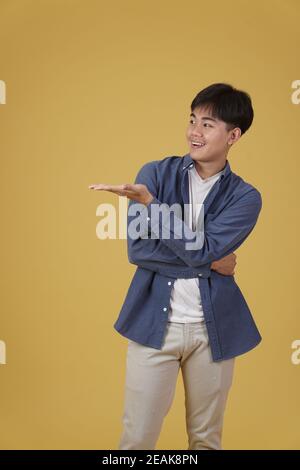 ritratto di felice bel giovane asiatico vestito casualmente presentante con gesto aperto della mano palma al copyspace isolato su giallo sfondo studio Foto Stock