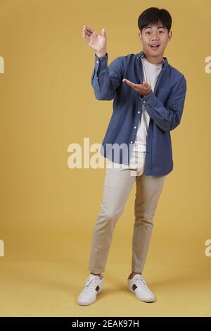 ritratto di felice bel giovane asiatico vestito casualmente presentante con gesto aperto della mano palma al copyspace isolato su giallo sfondo studio Foto Stock