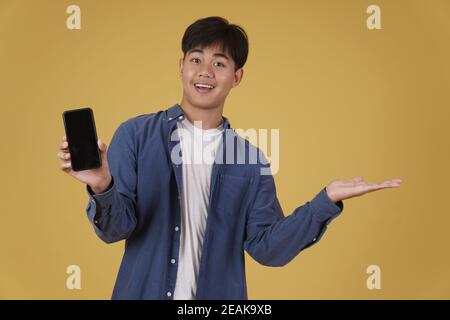 ritratto di felice giovane asiatico vestito casualmente sorridente mostrando schermo bianco vuoto dello smartphone isolato su sfondo giallo dello studio Foto Stock