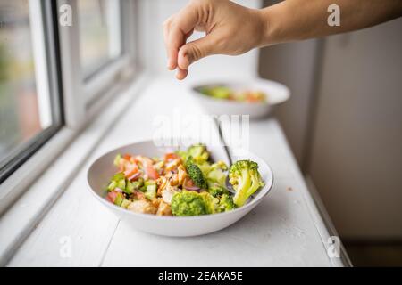 Mano aggiungendo un pizzico di sale ad un broccoli e. insalata di pollo accanto a una finestra Foto Stock