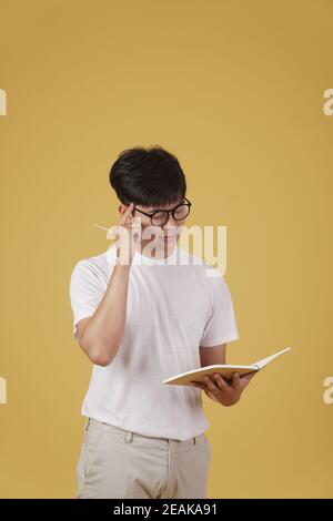 premuroso giovane studente asiatico vestito casualmente occhiali tenendo notebook pensiero isolato su sfondo giallo studio Foto Stock