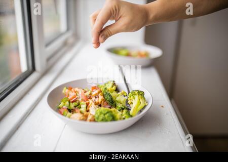 Mano aggiungendo un pizzico di sale ad un broccoli e. insalata di pollo accanto a una finestra Foto Stock