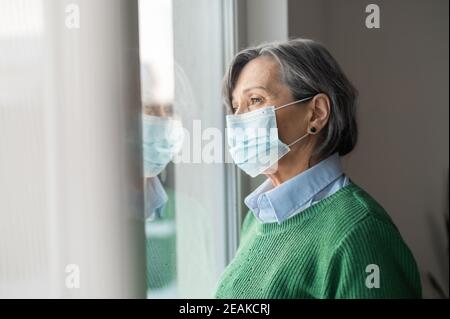 Una donna anziana con capelli grigi di media età che indossa un pullover verde e una protezione maschera facciale guardando la finestra e sentendosi triste depresso e solo Foto Stock