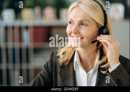 Ritratto di un'affascinante donna d'affari di mezza età con un abito elegante e una cuffia, seduto alla scrivania. Amichevole femmina lavora come operatore di call center, consulenza clienti, lavorando nel servizio di supporto Foto Stock