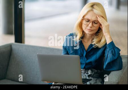Donna elegante matura in occhiali si siede sul divano, con mal di testa. Esausta donna caucasica di affari sta usando il laptop, ritenendosi stanco dopo il lavoro in linea, concetto di overwork Foto Stock