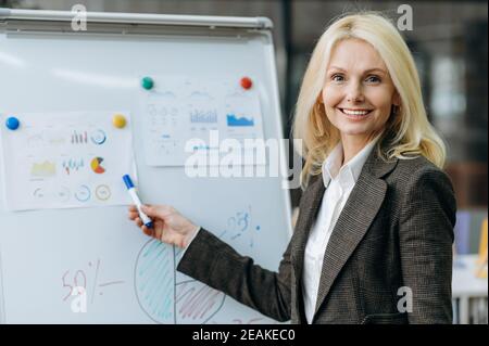 La signora di affari overjoyed fa la presentazione alla conferenza in linea. Bella donna anziana in abbigliamento formale guarda la fotocamera, sorridendo. Elegante donna si trova vicino alla lavagna a fogli mobili in un ufficio moderno Foto Stock