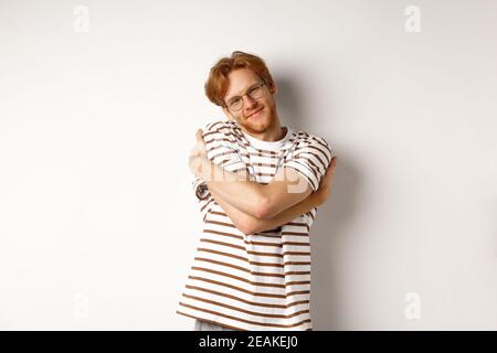 Bell'uomo sorridente con i capelli rossi e la barba che si rilassa, abbracciandosi e fissando il volto felice della fotocamera, sfondo bianco Foto Stock