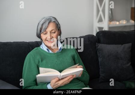 Nonna felice anziana donna matura in un maglione verde, seduta sul divano e leggendo un libro, donna di mezza età godendo il suo tempo libero in Foto Stock