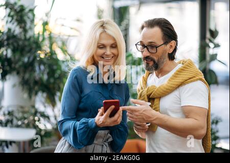 Soddisfatti colleghi caucasici maturi vestiti con stile, in piedi in un ufficio moderno, utilizzando smartphone, navigando in internet. Uomo anziano e donna elegante prendere una pausa dal lavoro, surf rete Foto Stock