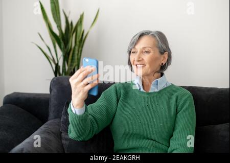 Donna matura ed eccitata che indossa un pullover verde seduto sul divano nel soggiorno, tenendo un telefono davanti al viso, facetiming il suo vecchio Foto Stock