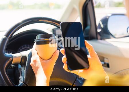 donna che beve caffè caldo takeaway tazza all'interno di una macchina e. tramite smartphone Foto Stock