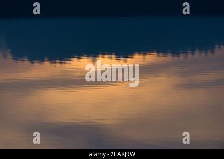 Concentratevi sull'acqua delicatamente increspata, riflettendo un treeline arancione al tramonto Foto Stock