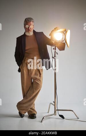 Foto a lunghezza intera di un uomo di mezza età con capelli grigi alla moda in occhiali che guardano la fotocamera, in piedi accanto al riflettore dello studio mentre si posa su sfondo bianco. Foto di moda, concetto di stile Foto Stock