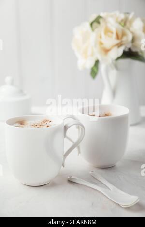 Due tazze di caffè in porcellana bianca. Bouquet di delicati fiori di crema sullo sfondo. Mattina romantica per due. Foto Stock