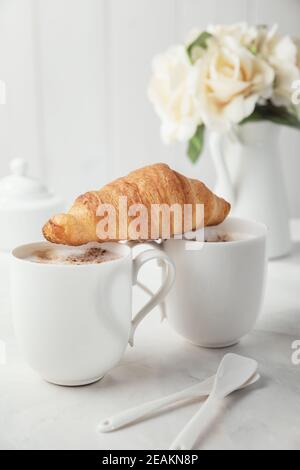 Due tazze bianche di caffè in porcellana con croissant. Bouquet di delicati fiori di crema sullo sfondo. Mattina romantica per due Foto Stock