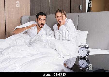 La coppia interracial stressata in accappatoi seduti sul letto vicino all'allarme orologio in primo piano sfocato in hotel Foto Stock