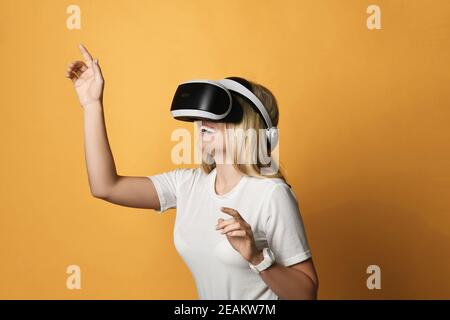 La donna bionda ridente in t-shirt utilizza gli occhiali VR funziona in realtà virtuale premendo il tasto con le dita. Vista laterale Foto Stock
