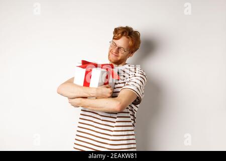 Concetto di giorno e vacanza di San Valentino. Felice uomo rosso abbracciando la sua confezione regalo e sorridendo felice, in piedi su sfondo bianco Foto Stock