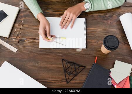 vista parziale dell'architetto afro-americano che tiene un divisorio vicino al libro di schizzo sulla scrivania a casa Foto Stock
