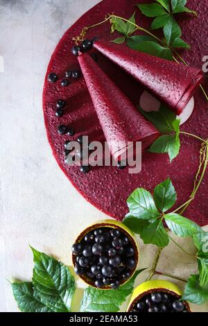 Pastila ribes nero su sfondo chiaro. La dolcezza viene arricciata in un cono. Nelle vicinanze si trova un ramo con foglie verdi. Ribes nero in un recipiente in ceramica. Foto verticale. Foto Stock