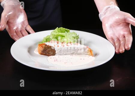 Le mani dello chef servono piatti da un pezzo di salmone in una cremosa salsa caviale su un piatto bianco con foglie di insalata. Foto macro. Foto Stock