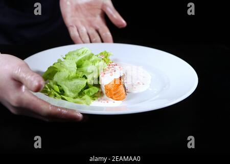 Salmone in cremosa salsa caviale su un piatto bianco. Il cuoco serve il piatto preparato. Foglie di lattuga. Foto su sfondo nero. Persona irriconoscibile. Foto Stock