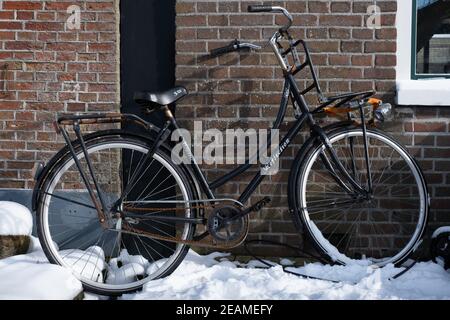 Bici delle Signore nere con catena arrugginita parcheggiata contro un mattone Muro nella neve nei Paesi Bassi Foto Stock